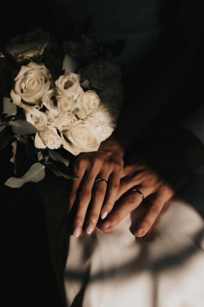Mani degli sposi con le fedi, in ombra, accanto a un mazzo di rose bianche